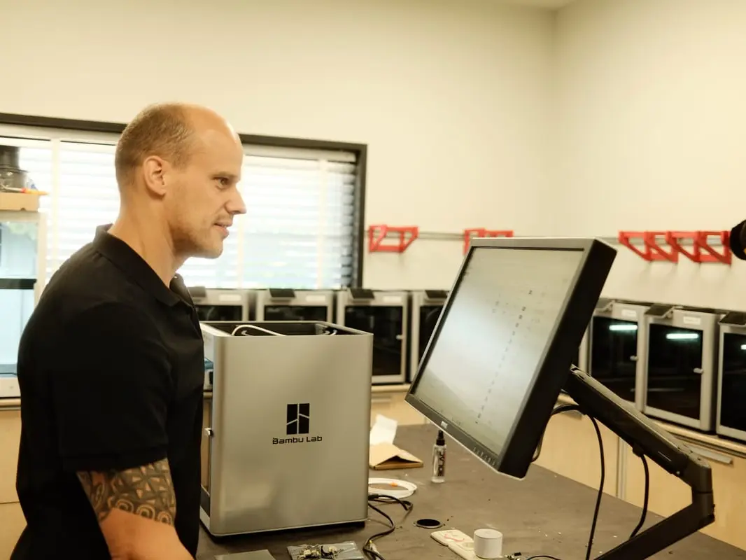 Ein Mitarbeiter kontrolliert über seinen Computer die Druckfarm, die im Hintergrund zu sehen ist.