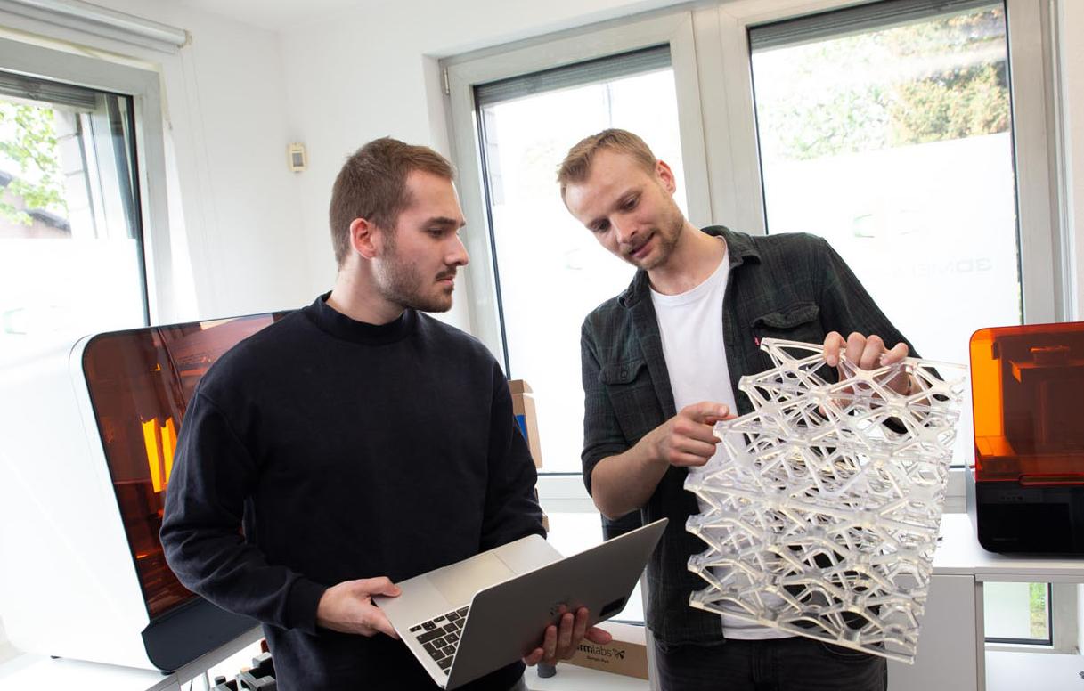 links person mit laptop in der hand, rechts person mit 3d druck erklärt der anderen person etwas