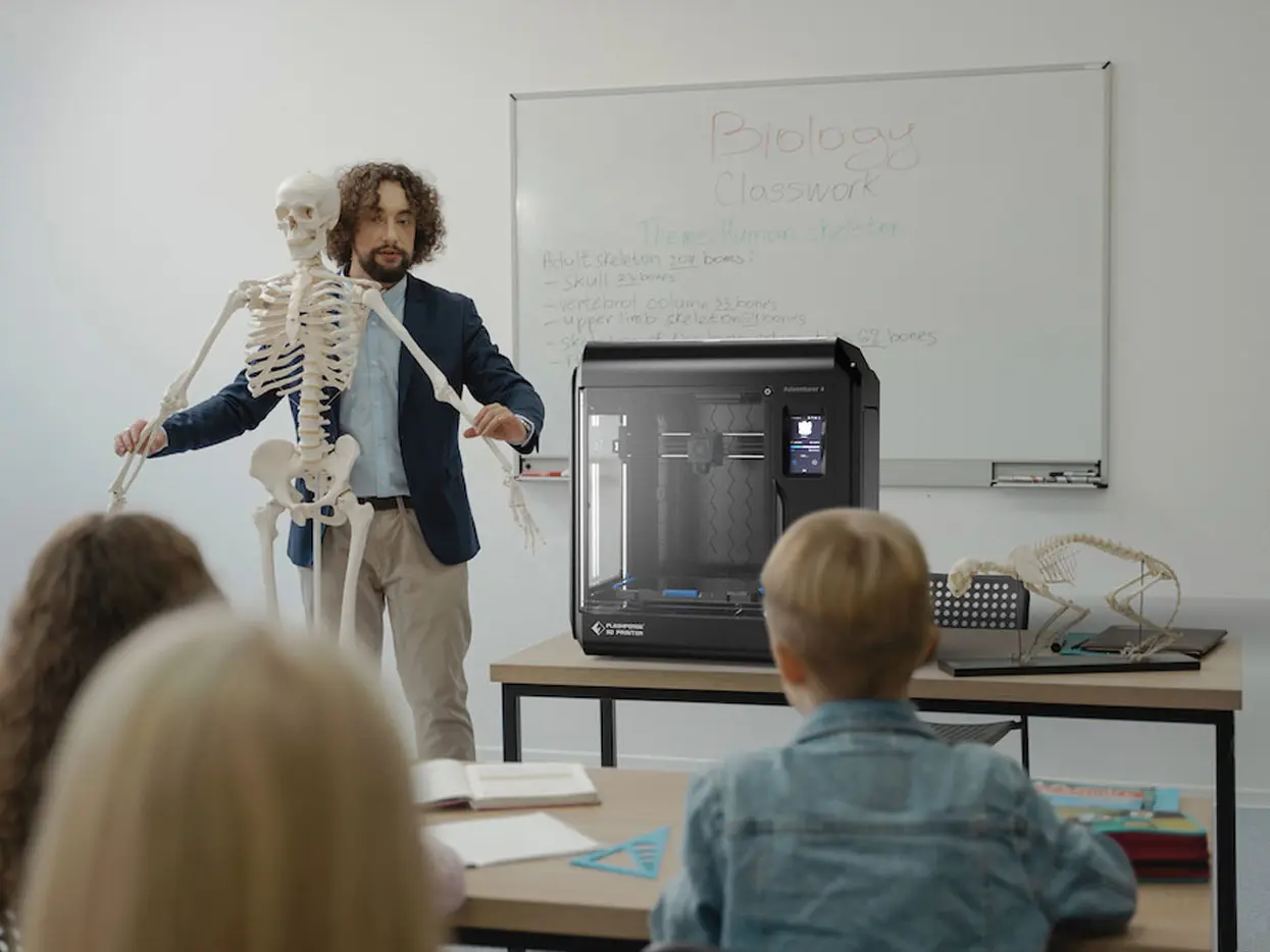 Eine Lehrkraft steht vor seinen Schülerinnen und auf seinem Tisch steht ein 3D Drucker